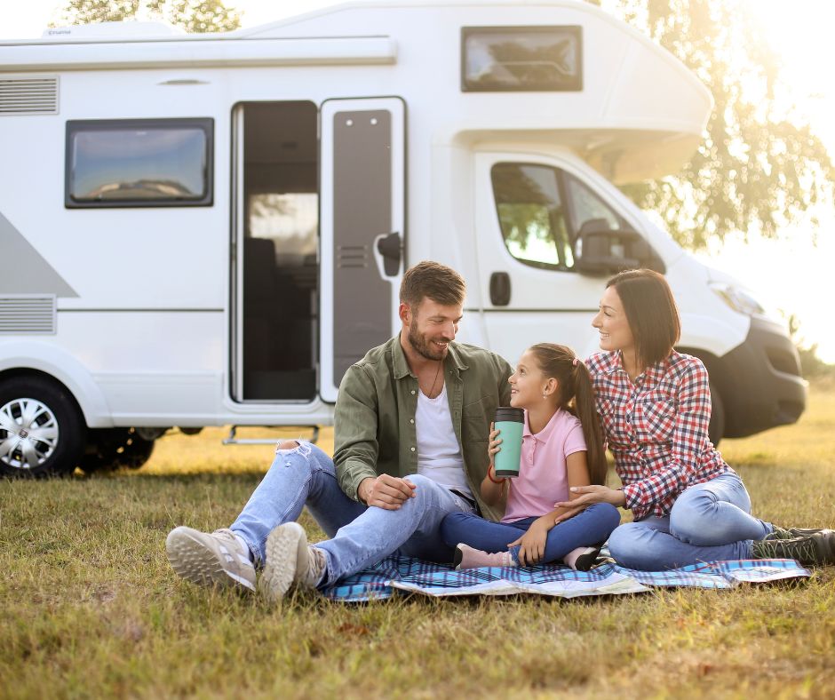 Two adults and a child on a picnic blanket in front of a camper