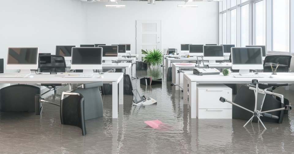 An office that is flooded with water. Some chairs, a printer, a plant and other items are floating in the water.