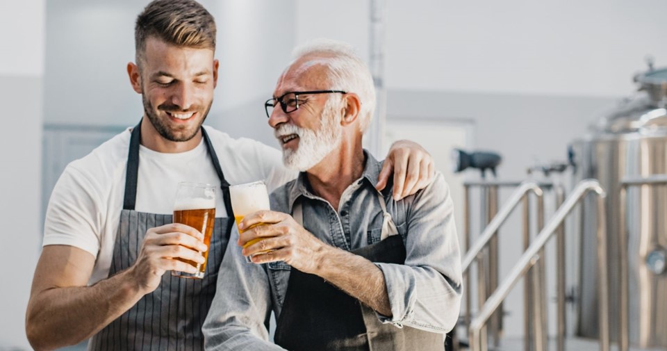 A young man cheersing beers with an older man in a brewery