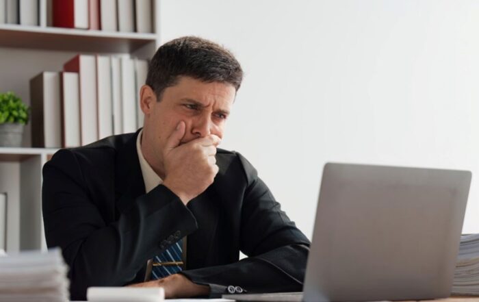 A man looking at his laptop in front of him while covering his mouth and looking concerned.