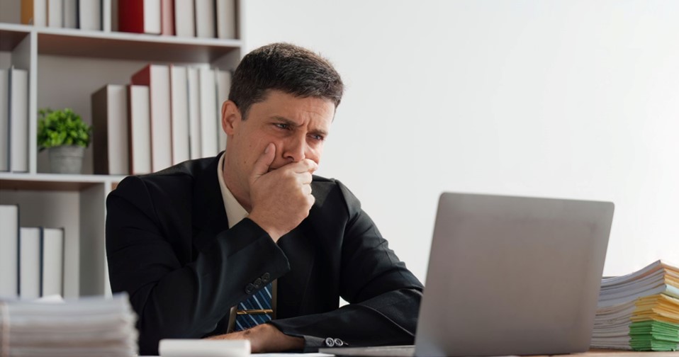 A man looking at his laptop in front of him while covering his mouth and looking concerned.