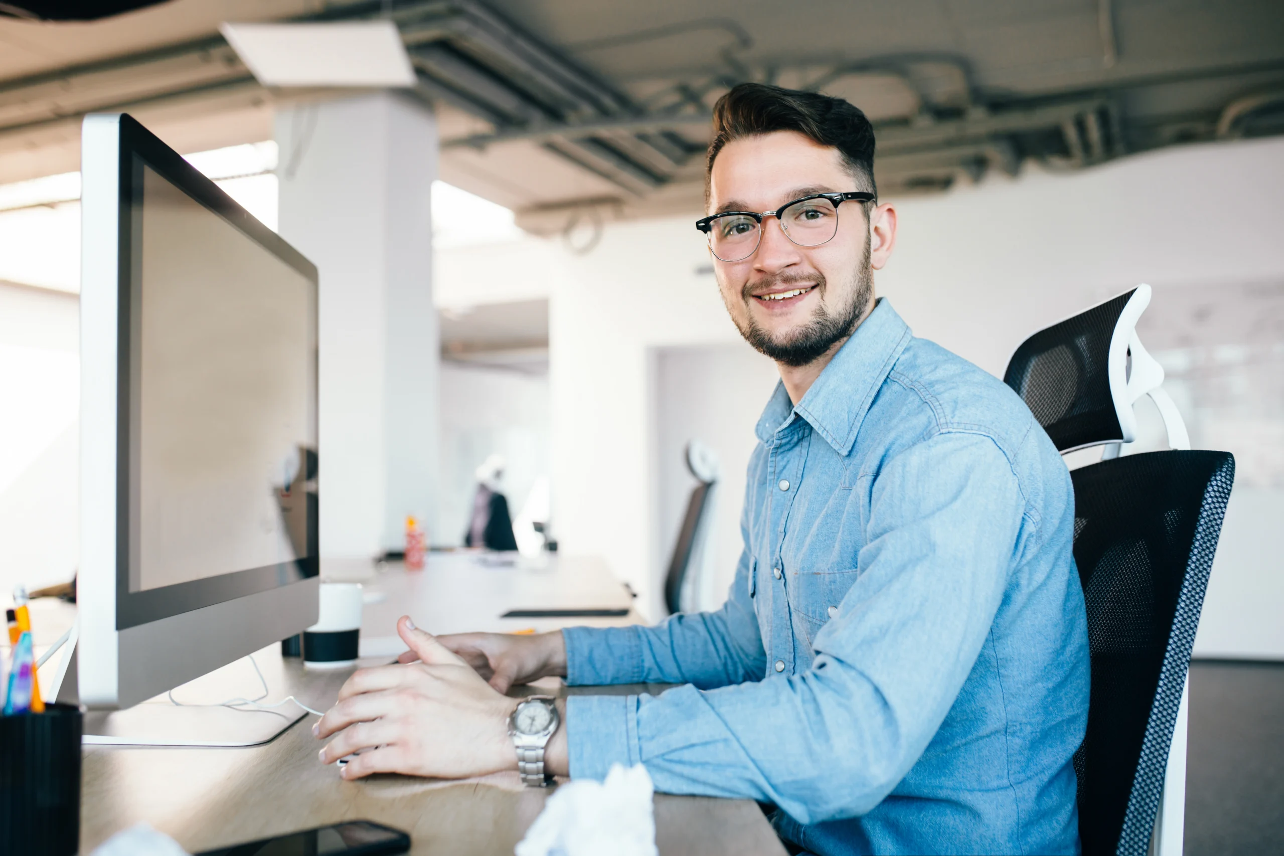Young dark-haired man in glasses is working with a computer at his desktop in office. He wears blue shirt and smiles to the camera. View from side